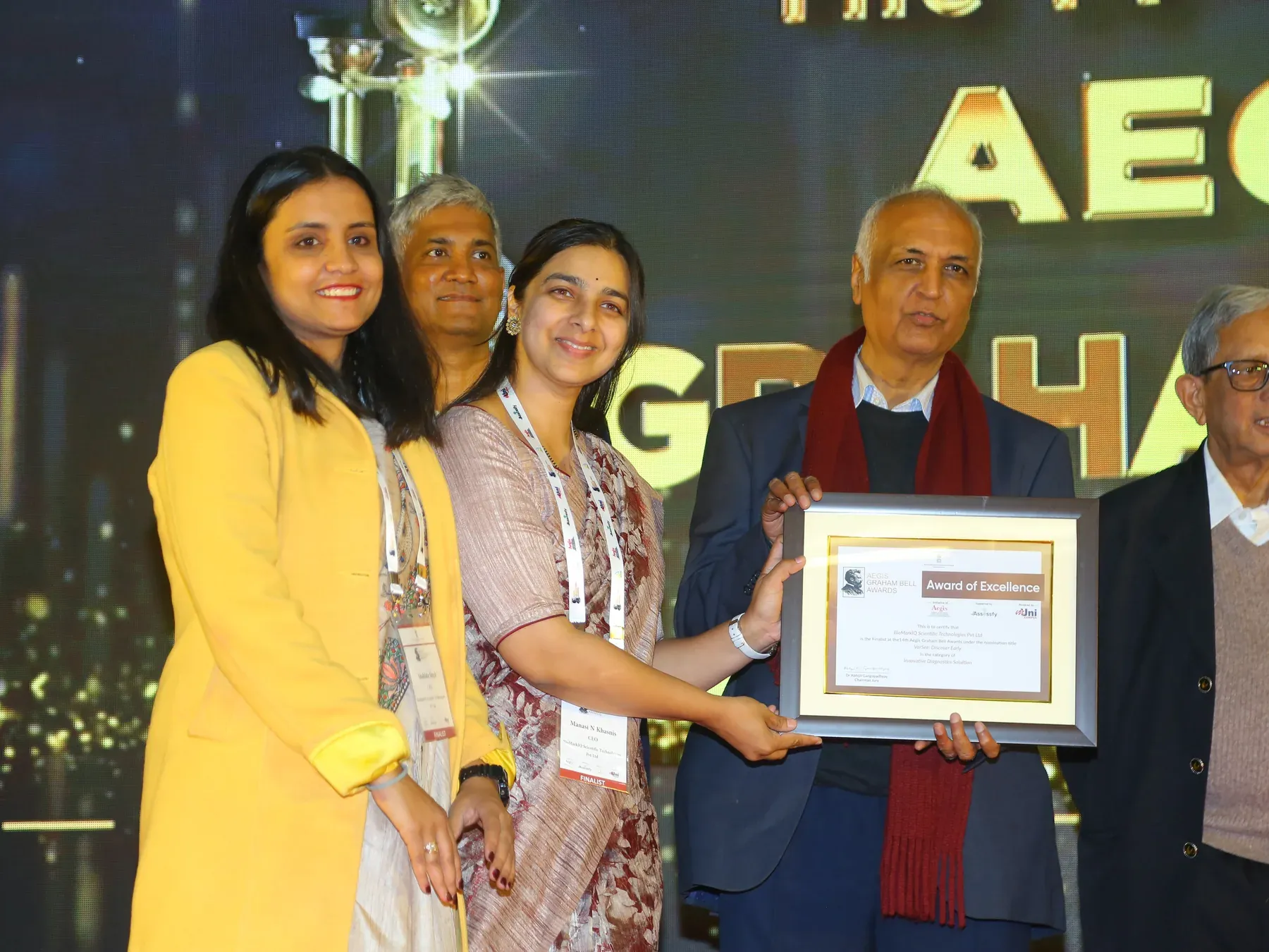 Manasi Khasnis and a colleague accept the Aegis Graham Bell Awards certificate of excellence on stage.