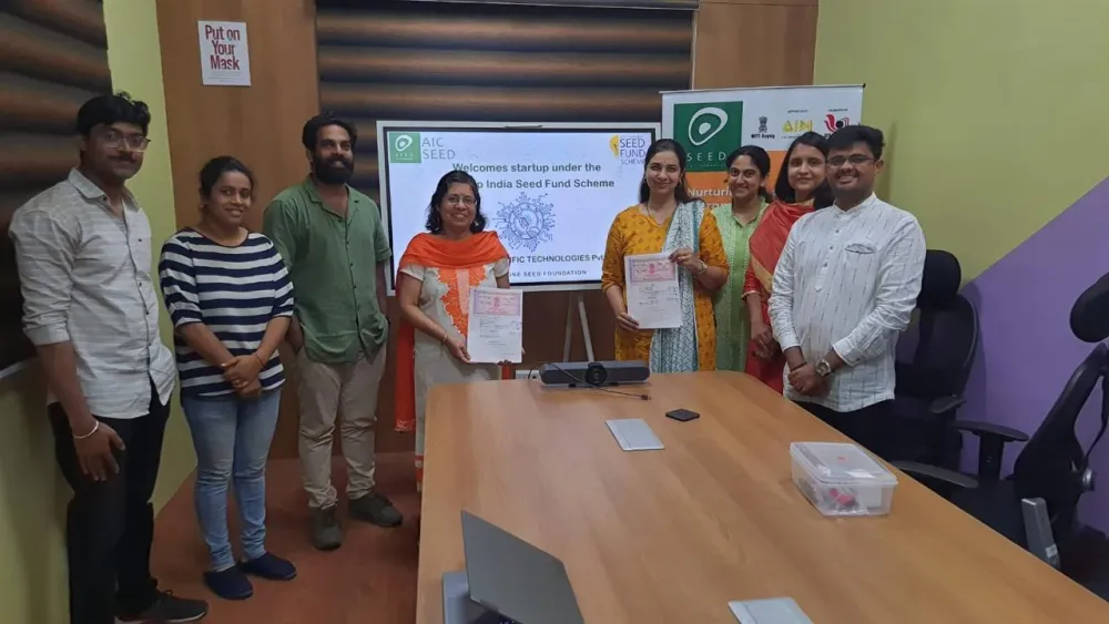 Eight people in a meeting room, two holding signed documents, before a screen welcoming the startup under the Startup India Seed Fund Scheme.