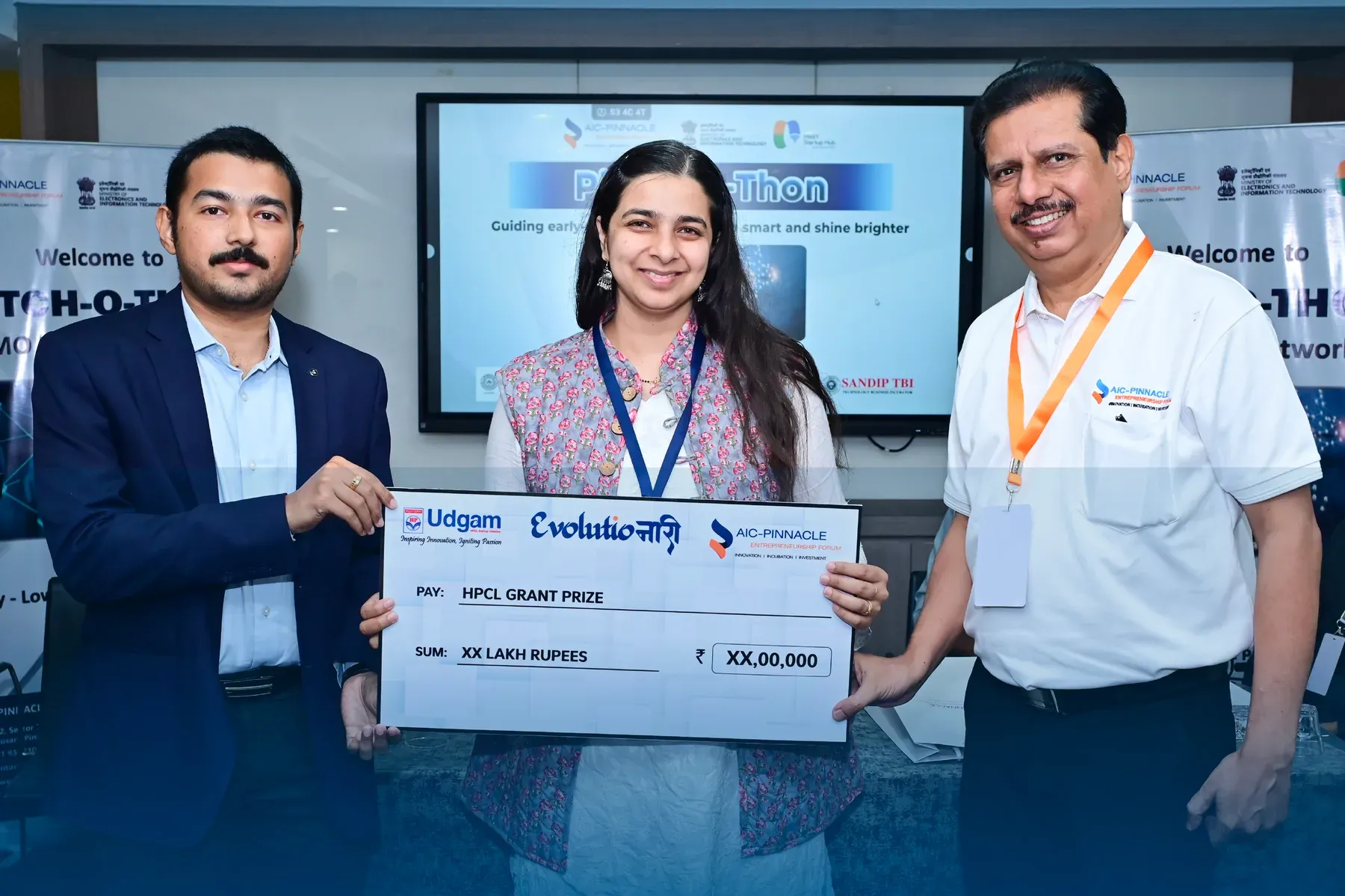 Manasi Khasnis holds an EvolutioNari HPCL grant prize cheque between two hosts at AIC-Pinnacle’s Pitch-o-Thon.