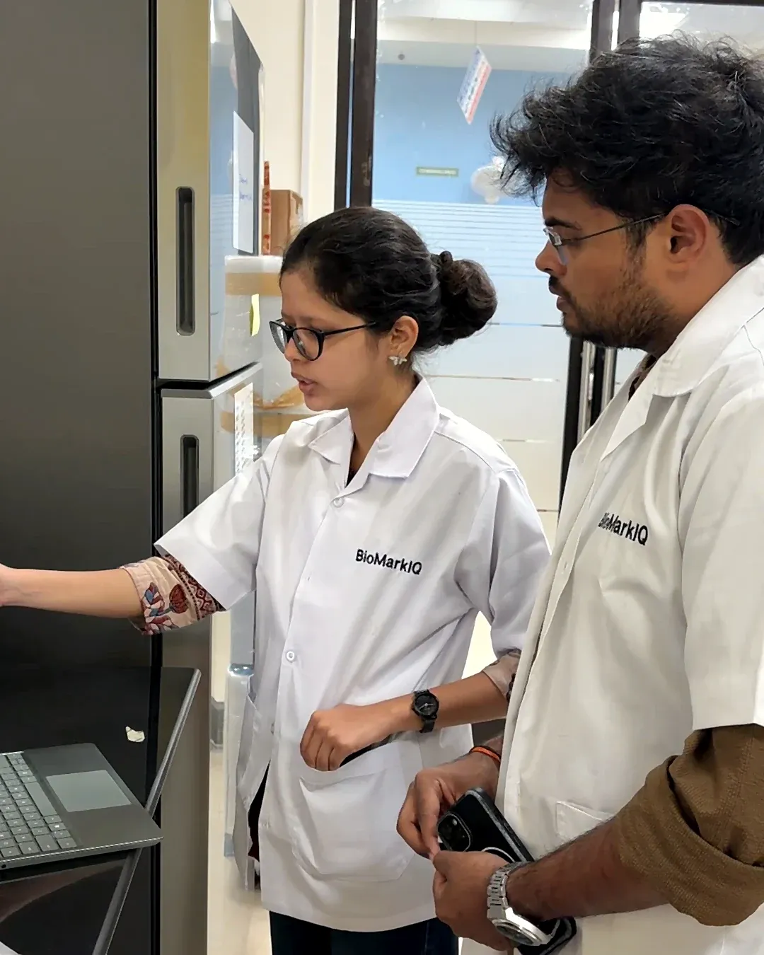 Two BioMarkIQ team members in lab coats at a laboratory analyser, one pointing at its screen.
