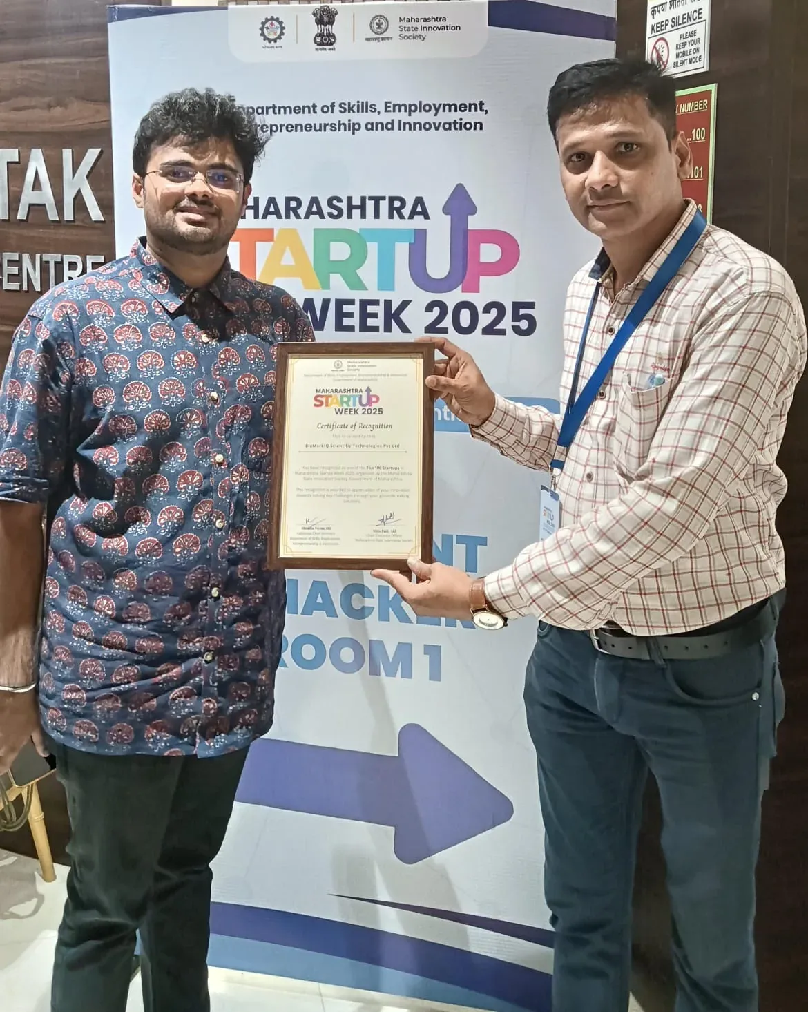 Two men hold BioMarkIQ’s Maharashtra Startup Week 2025 Certificate of Recognition in front of the event banner.