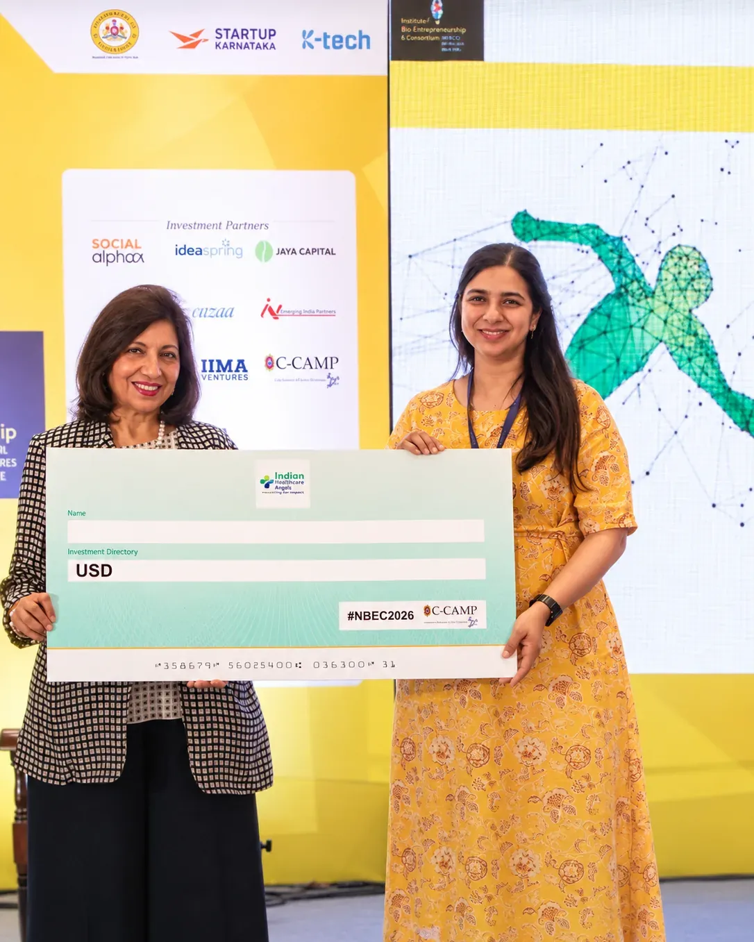 Manasi Khasnis on stage at NBEC 2026, holding an Indian Healthcare Angels cheque.
