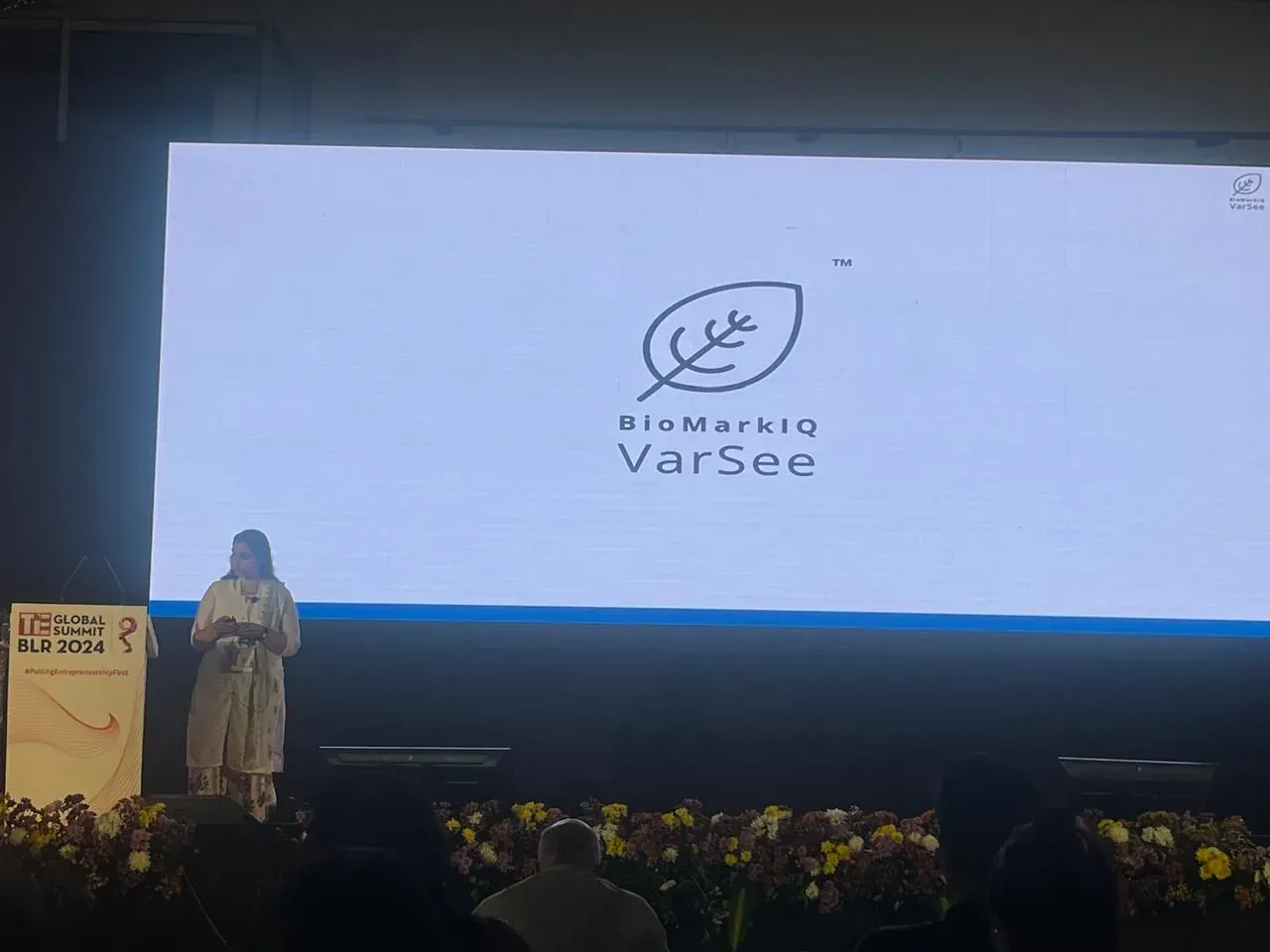 A BioMarkIQ founder presents at TiE Global Summit BLR 2024, the BioMarkIQ VarSee leaf logo filling the screen behind her.
